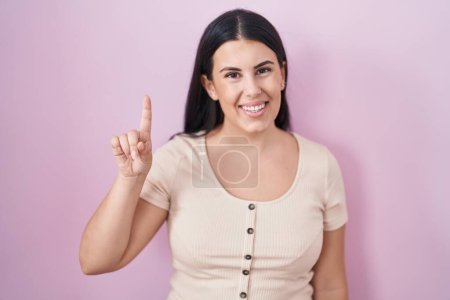 Téléchargez les photos : Jeune femme hispanique debout sur fond rose montrant et pointant vers le haut avec le doigt numéro un tout en souriant confiant et heureux. - en image libre de droit