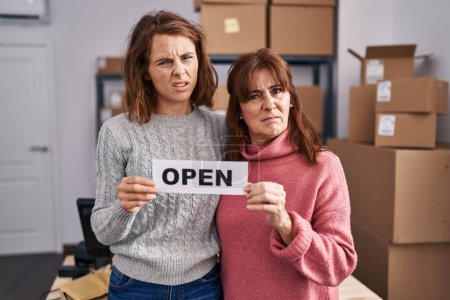 Foto de Two women working at small business ecommerce holding open banner clueless and confused expression. doubt concept. - Imagen libre de derechos