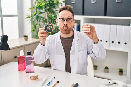 Photo for Middle age caucasian dentist man holding denture and doughnuts making fish face with mouth and squinting eyes, crazy and comical. - Royalty Free Image