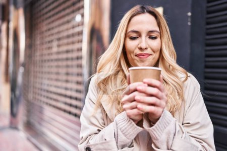Téléchargez les photos : Jeune femme blonde souriante boire du café en toute confiance à la rue - en image libre de droit