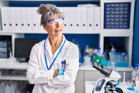 Téléchargez les photos : Middle age woman scientist sitting with arms crossed gesture at laboratory - en image libre de droit