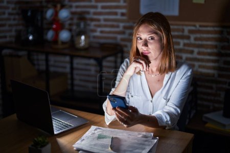 Photo for Young redhead woman working at the office at night serious face thinking about question with hand on chin, thoughtful about confusing idea - Royalty Free Image