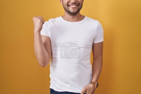 Photo for Hispanic man wearing white t shirt over yellow background screaming proud, celebrating victory and success very excited with raised arms - Royalty Free Image