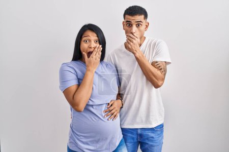 Foto de Young hispanic couple expecting a baby standing over background shocked covering mouth with hands for mistake. secret concept. - Imagen libre de derechos