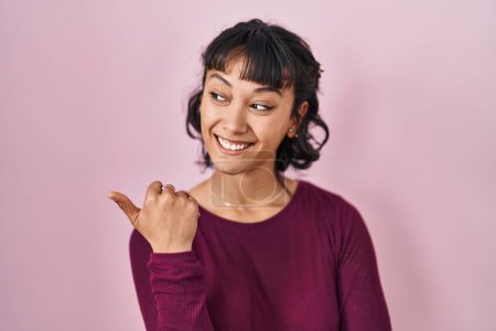 Foto de Young beautiful woman standing over pink background smiling with happy face looking and pointing to the side with thumb up. - Imagen libre de derechos