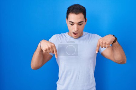 Photo for Young hispanic man standing over blue background pointing down with fingers showing advertisement, surprised face and open mouth - Royalty Free Image