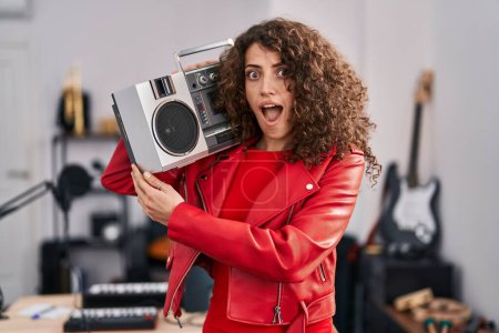 Téléchargez les photos : Hispanic woman with curly hair holding boombox, listening to music scared and amazed with open mouth for surprise, disbelief face - en image libre de droit