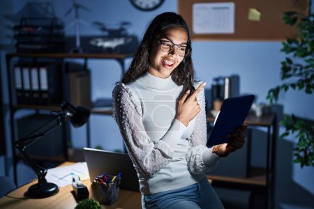 Téléchargez les photos : Jeune femme brésilienne utilisant le pavé tactile la nuit travaillant au bureau avec un grand sourire sur le visage, pointant avec le doigt de la main vers le côté regardant la caméra. - en image libre de droit
