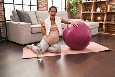 Foto de Joven embarazada sentada en esterilla de yoga con pelota de pilates celebrando la victoria con sonrisa feliz y expresión ganadora con las manos levantadas - Imagen libre de derechos