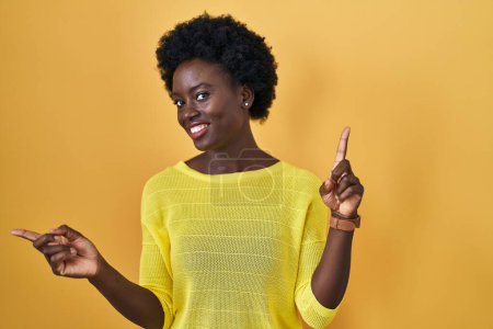 Téléchargez les photos : Jeune femme africaine debout sur studio jaune souriant confiant pointant avec les doigts vers différentes directions. espace de copie pour la publicité - en image libre de droit