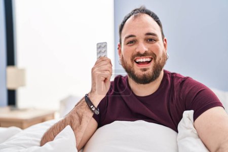 Foto de Hombre hispano de talla grande con barba en la cama sosteniendo píldoras que parecen positivas y felices de pie y sonriendo con una sonrisa confiada mostrando los dientes - Imagen libre de derechos