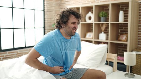 Téléchargez les photos : Young hispanic man suffering for stomach ache sitting on bed at bedroom - en image libre de droit