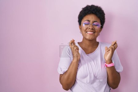 Téléchargez les photos : Jeune femme afro-américaine debout sur fond rose geste doigt croisé souriant avec espoir et les yeux fermés. chance et concept superstitieux. - en image libre de droit