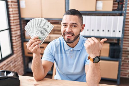 Téléchargez les photos : Hispanique homme travaillant à la petite entreprise commerce électronique tenant des dollars pointant pouce vers le côté souriant heureux avec la bouche ouverte - en image libre de droit