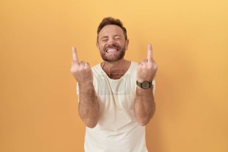 Foto de Middle age man with beard standing over yellow background showing middle finger doing fuck you bad expression, provocation and rude attitude. screaming excited - Imagen libre de derechos
