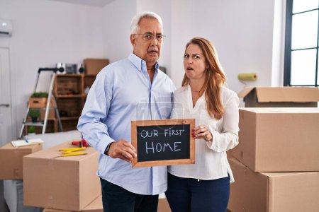 Téléchargez les photos : Middle age hispanic couple moving to a new home holding banner in shock face, looking skeptical and sarcastic, surprised with open mouth - en image libre de droit