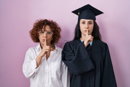 Foto de Hispanic mother and daughter wearing graduation cap and ceremony robe asking to be quiet with finger on lips. silence and secret concept. - Imagen libre de derechos