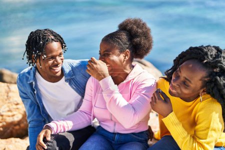 Foto de Amigos afroamericanos sonriendo confiados sentados en la roca juntos a la orilla del mar - Imagen libre de derechos