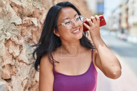 Foto de Young chinese woman smiling confident listening audio message by the smartphone at street - Imagen libre de derechos