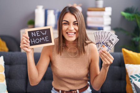 Photo for Young hispanic woman holding blackboard with new home text and dollars smiling and laughing hard out loud because funny crazy joke. - Royalty Free Image