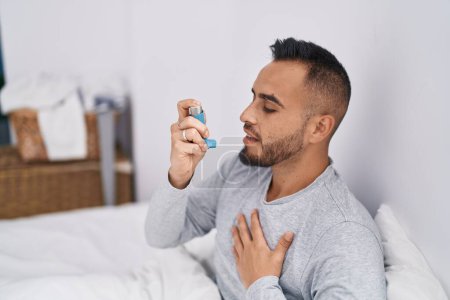 Photo for Young hispanic man using inhaler sitting on bed at bedroom - Royalty Free Image