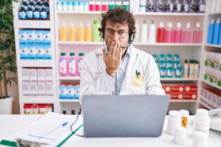 Photo for Hispanic young man working at pharmacy drugstore working with laptop covering mouth with hand, shocked and afraid for mistake. surprised expression - Royalty Free Image