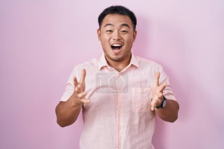 Téléchargez les photos : Jeune homme chinois debout sur fond rose célébrant fou et étonné pour le succès avec les bras levés et les yeux ouverts hurlant excité. concept gagnant - en image libre de droit