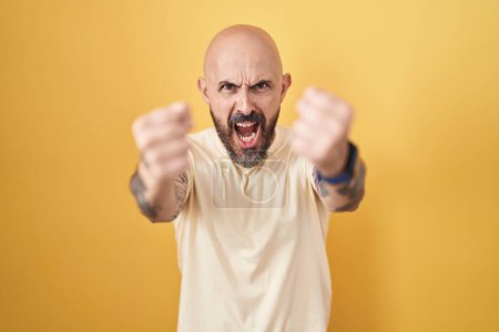 Téléchargez les photos : Homme hispanique avec des tatouages debout sur fond jaune en colère et fou levant les poings frustrés et furieux tout en criant de colère. rage et concept agressif. - en image libre de droit