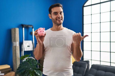 Photo for Young hispanic man with beard holding piggy bank at new home pointing thumb up to the side smiling happy with open mouth - Royalty Free Image