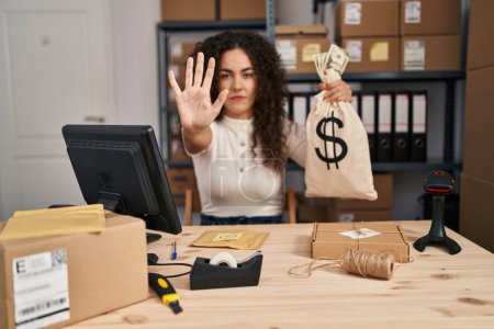 Photo for Young hispanic woman working at small business ecommerce holding money bag with open hand doing stop sign with serious and confident expression, defense gesture - Royalty Free Image