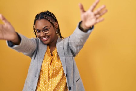 Téléchargez les photos : Femme afro-américaine avec des tresses debout sur fond jaune regardant la caméra souriant à bras ouverts pour étreindre. expression joyeuse embrassant le bonheur. - en image libre de droit