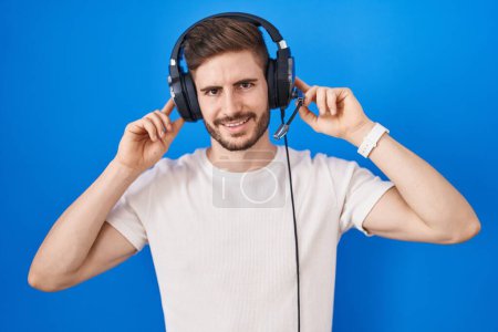 Foto de Hispanic man with beard listening to music wearing headphones smiling pulling ears with fingers, funny gesture. audition problem - Imagen libre de derechos