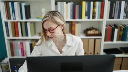 Foto de Young blonde woman student using computer writing on notebook at library university - Imagen libre de derechos