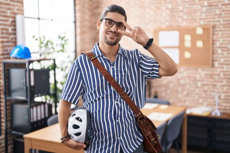Photo for Hispanic man with long hair working at the office holding bike helmet doing peace symbol with fingers over face, smiling cheerful showing victory - Royalty Free Image