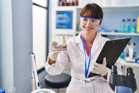 Photo for Young beautiful hispanic woman scientist holding clipboard and bottle at pharmacy - Royalty Free Image