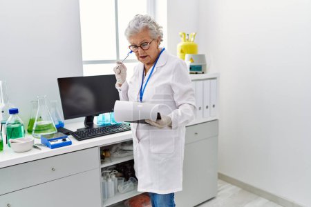 Téléchargez les photos : Femme âgée aux cheveux gris portant un uniforme de scientifique avec une expression de doute au laboratoire - en image libre de droit