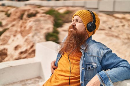 Téléchargez les photos : Jeune rousse homme écouter de la musique assis sur le banc à la rue - en image libre de droit