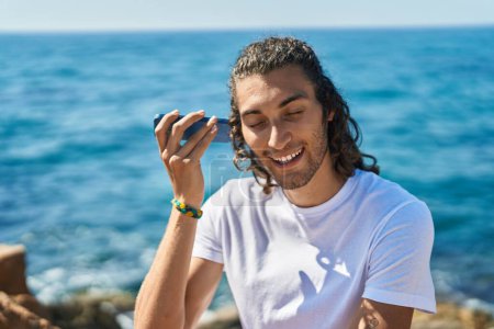 Téléchargez les photos : Young hispanic man listening audio message by the smartphone sitting on rock at seaside - en image libre de droit