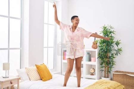 Téléchargez les photos : Femme afro-américaine souriante danse confiante sur le lit dans la chambre - en image libre de droit