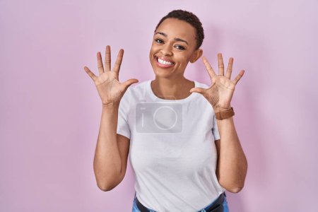 Foto de Hermosa mujer afroamericana de pie sobre fondo rosa mostrando y apuntando hacia arriba con los dedos número diez mientras sonríe confiado y feliz. - Imagen libre de derechos