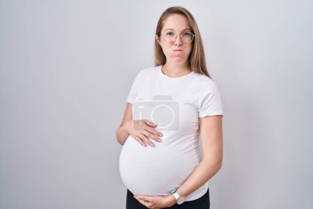 Téléchargez les photos : Young pregnant woman expecting a baby, touching pregnant belly puffing cheeks with funny face. mouth inflated with air, catching air. - en image libre de droit