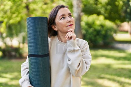 Téléchargez les photos : Middle age hispanic woman holding yoga mat at the park serious face thinking about question with hand on chin, thoughtful about confusing idea - en image libre de droit