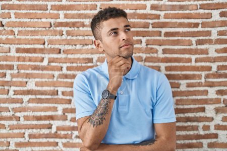 Photo for Brazilian young man standing over brick wall with hand on chin thinking about question, pensive expression. smiling with thoughtful face. doubt concept. - Royalty Free Image