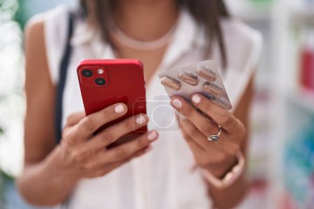 Téléchargez les photos : Jeune belle cliente hispanique utilisant un smartphone tenant des comprimés à la pharmacie - en image libre de droit