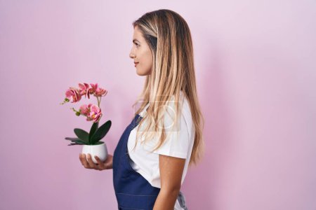 Foto de Mujer rubia joven con delantal de jardinero sosteniendo planta mirando a un lado, pose de perfil relajante con rostro natural y sonrisa confiada. - Imagen libre de derechos