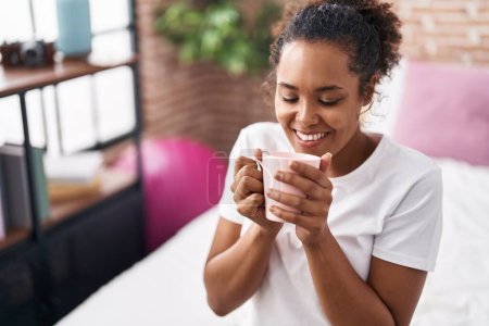 Téléchargez les photos : Femme afro-américaine buvant une tasse de café assise sur le lit dans la chambre - en image libre de droit