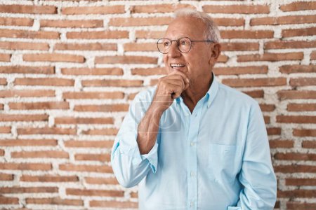 Photo for Senior man with grey hair standing over bricks wall with hand on chin thinking about question, pensive expression. smiling and thoughtful face. doubt concept. - Royalty Free Image