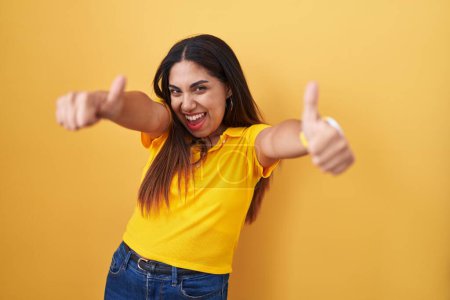 Téléchargez les photos : Young arab woman standing over yellow background approving doing positive gesture with hand, thumbs up smiling and happy for success. winner gesture. - en image libre de droit