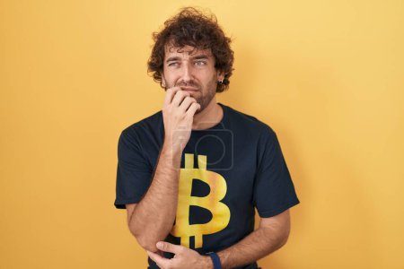 Foto de Hispanic young man wearing bitcoin t shirt looking stressed and nervous with hands on mouth biting nails. anxiety problem. - Imagen libre de derechos