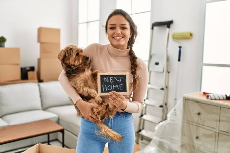 Photo for Young beautiful hispanic woman hugging dog holding blackboard at new home - Royalty Free Image